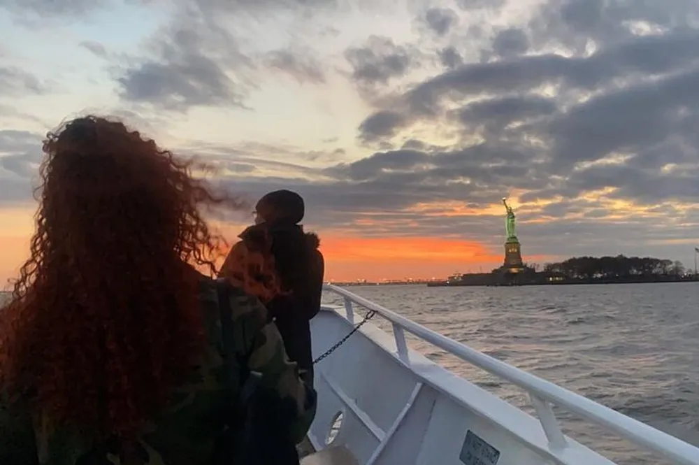 A person with curly red hair is looking towards the Statue of Liberty from a boat at sunset