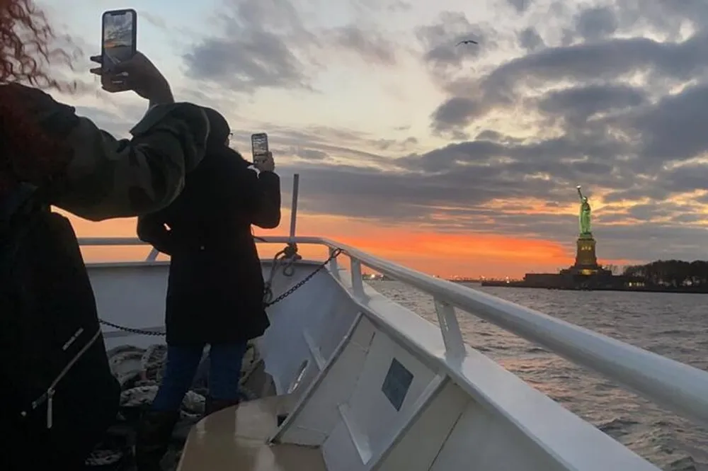 Two individuals are capturing the sunset and the Statue of Liberty with their smartphones from the deck of a boat