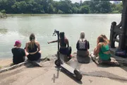 Four individuals are sitting by a lake in a park, with one pointing towards the water, while an electric scooter rests on the ground beside them.