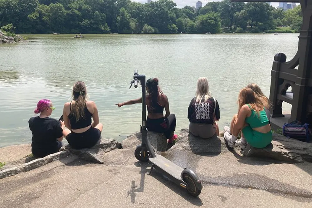 Four individuals are sitting by a lake in a park with one pointing towards the water while an electric scooter rests on the ground beside them