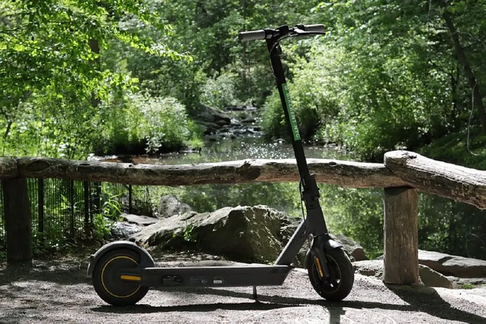 An electric scooter stands on a pathway in a serene forest setting with a small stream and lush greenery in the background