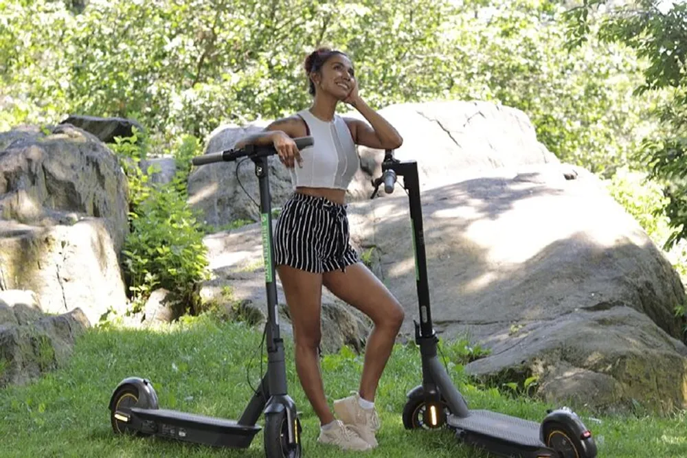 A woman is standing outdoors with a bright smile leaning on an electric scooter in a sunny park setting