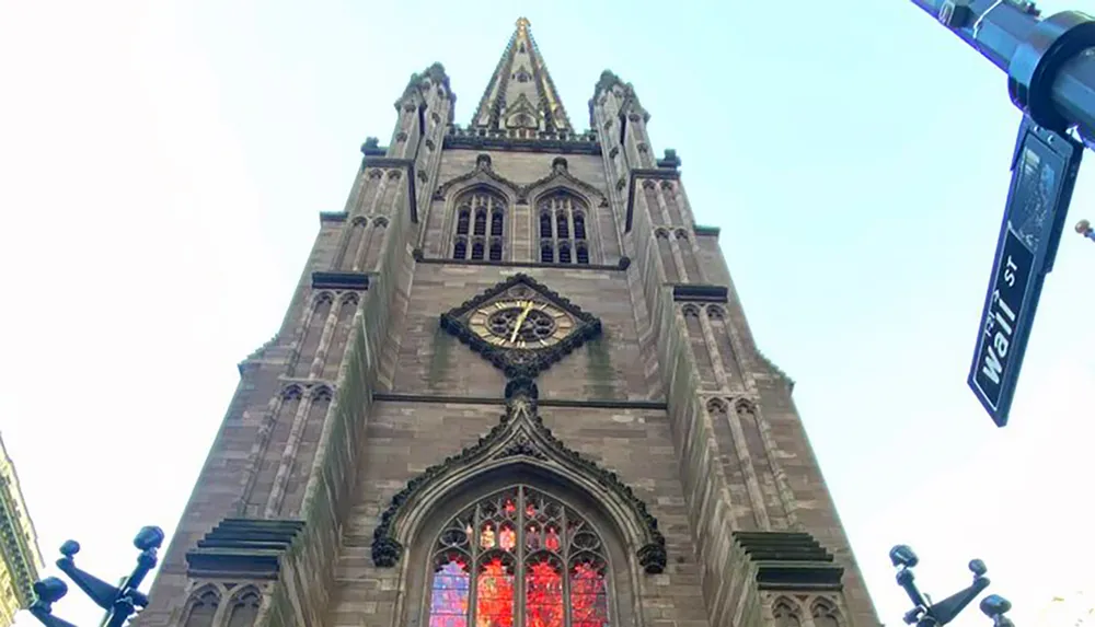 The image shows the towering Gothic architecture of a church facade with a large stained glass window set against a bright sky with a street sign indicating Wall St prominently in the foreground to the right