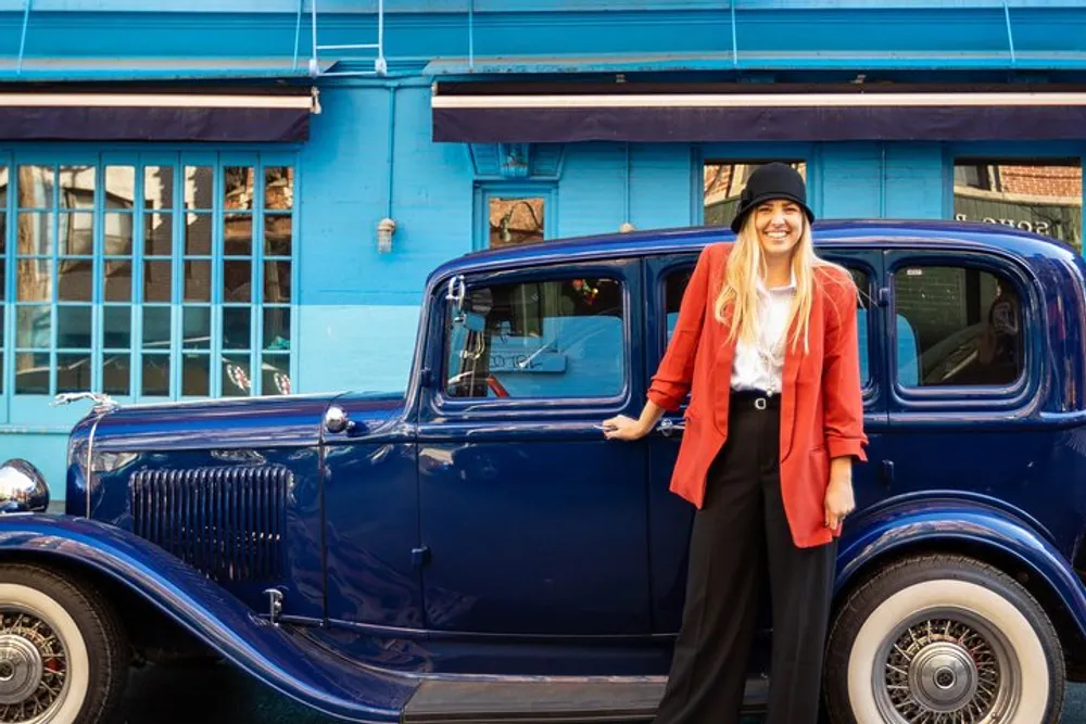 A smiling person in a red jacket and black hat stands next to a classic blue car in front of a building with a bright blue facade