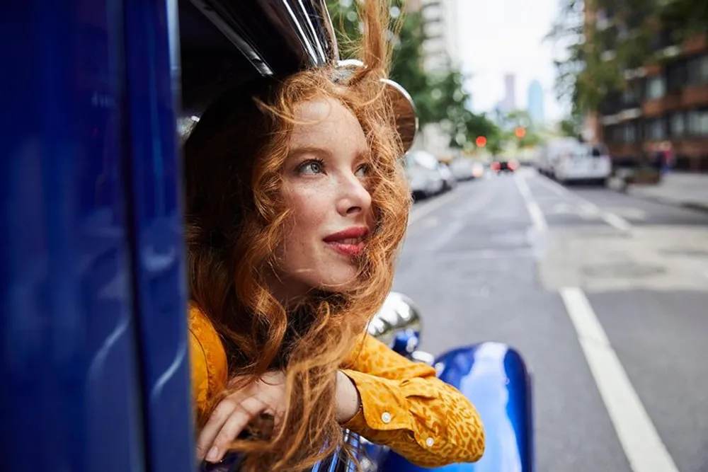 A woman with red hair is gazing out of the passenger window of a blue vehicle with a thoughtful expression on a tree-lined city street