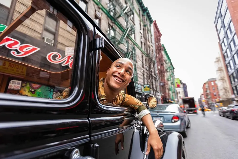 A happy person is leaning out of the passenger window of a vintage car smiling as they travel down an urban street lined with buildings and various signs