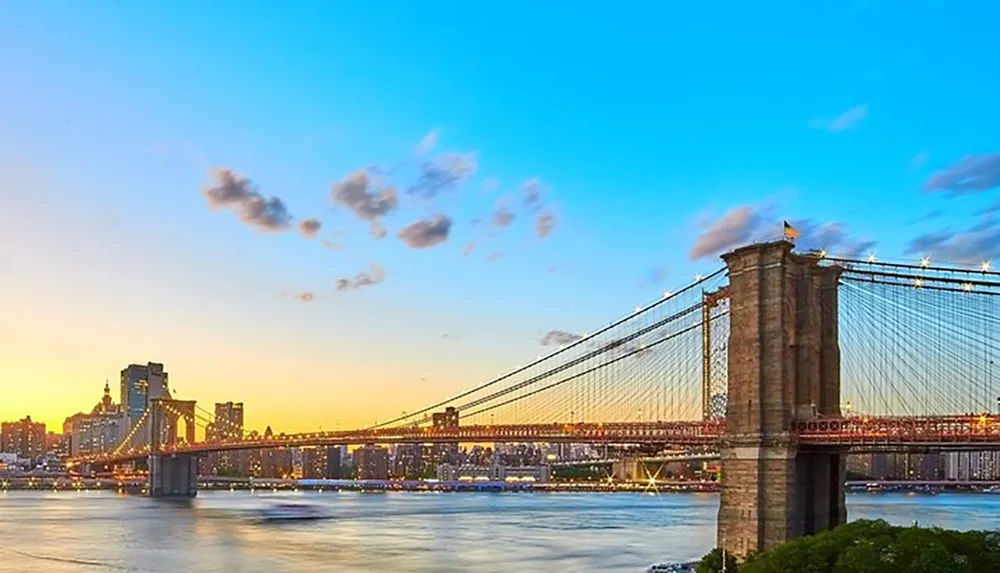 The image shows the Brooklyn Bridge spanning the East River during sunset with a vibrant sky backdrop in New York City