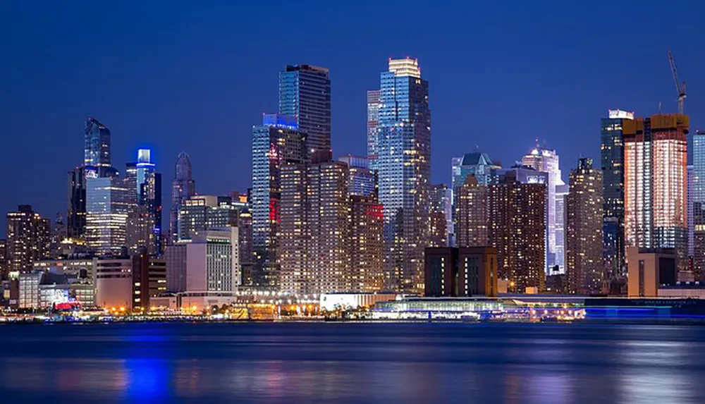 The image shows a vibrant city skyline at twilight with illuminated skyscrapers reflecting on the calm water in the foreground