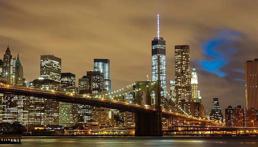 The image shows a nighttime view of the Brooklyn Bridge with the illuminated Manhattan skyline in the background