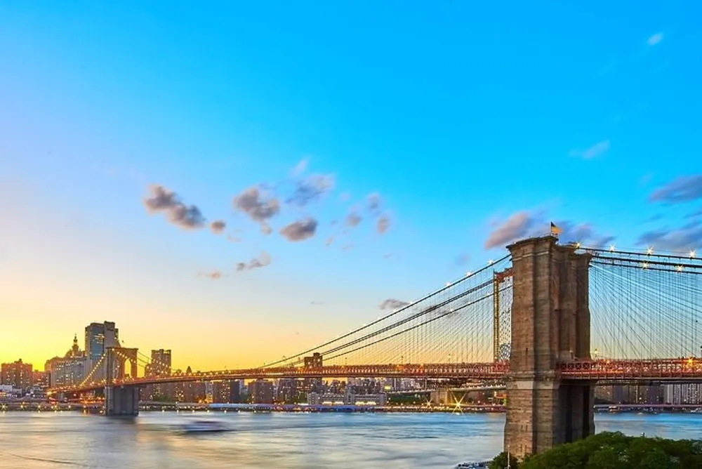 The Brooklyn Bridge stands majestically against a vibrant sunrise with a clear blue sky scattered with small clouds