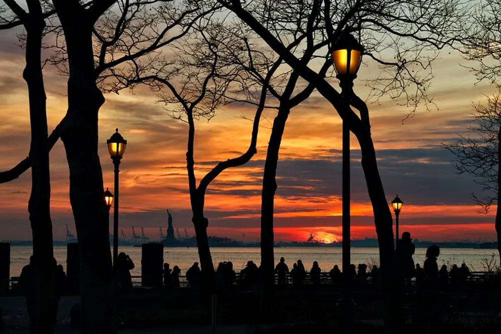 Silhouetted figures and bare trees are set against a vivid sunset sky with the glow of street lamps adding to the tranquil evening ambiance by the water