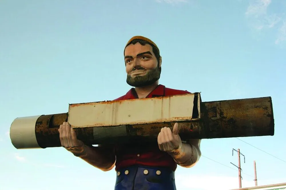 A large colorful statue of a bearded man in a cap and rolled-up sleeves carries an oversized cylindrical object on his shoulder against a sky backdrop