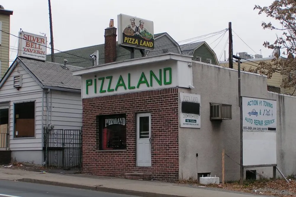 The image shows a small brick building with a prominent green PIZZALAND sign located on a street corner with adjacent businesses and signage