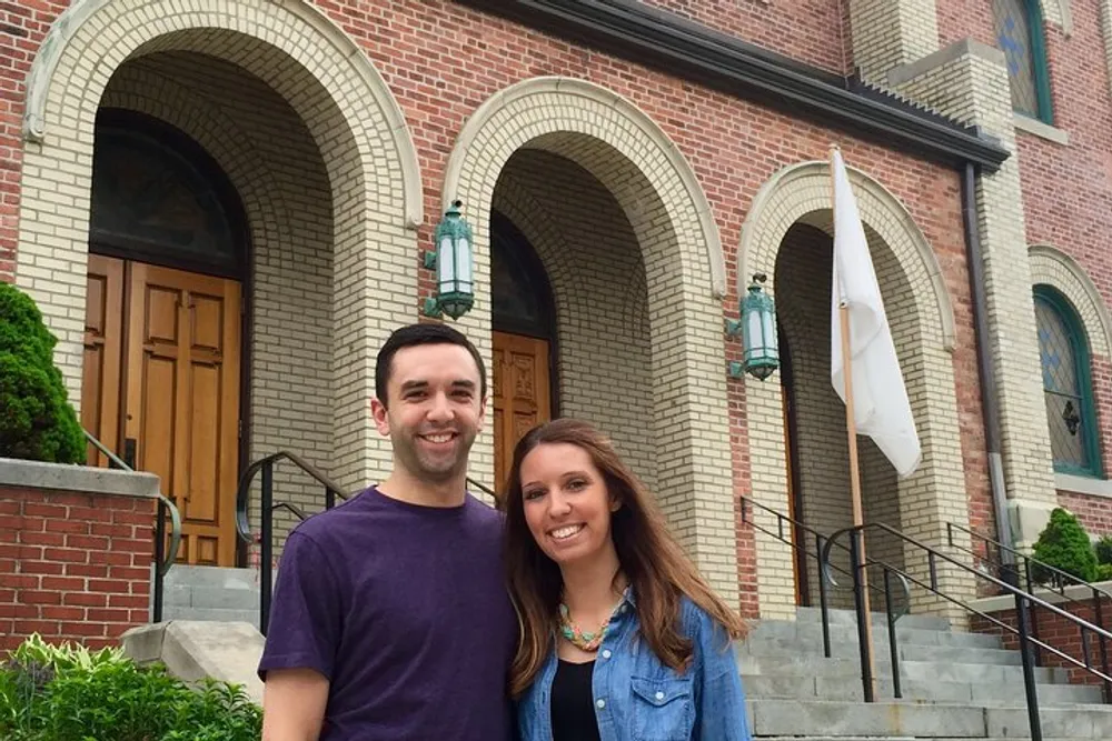Two people are smiling in front of a building with brick walls and arched entrance doors