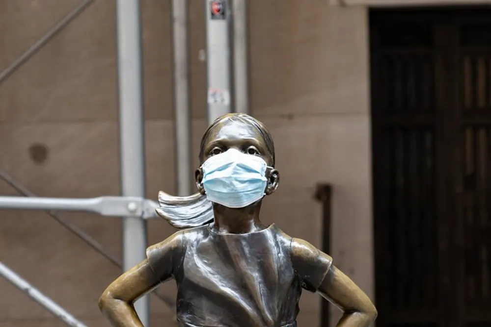 The image shows a bronze sculpture of a defiant young girl wearing a disposable face mask positioned against an urban backdrop with scaffolding