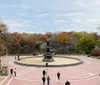 The image shows people walking around a large fountain with an angel statue on top set within an urban park featuring vibrant autumn foliage on the trees in the background