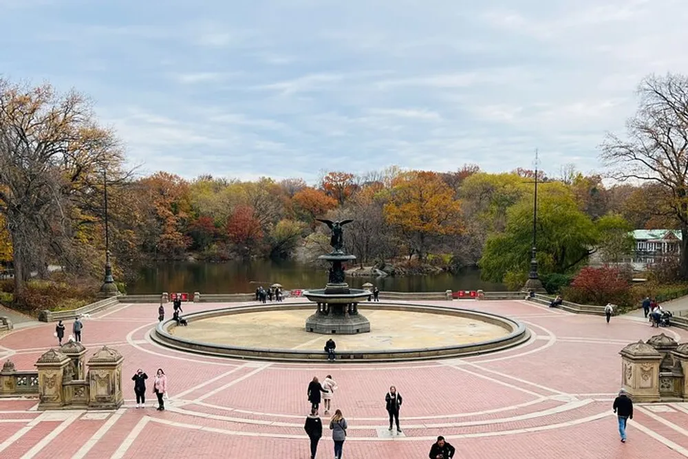 The image shows people walking around a large fountain with an angel statue on top set within an urban park featuring vibrant autumn foliage on the trees in the background