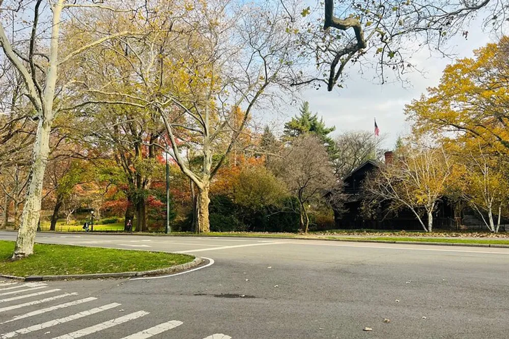 The image shows a peaceful autumn scene in a park with leafy trees displaying fall colors a curving road and a dark-colored building in the background under a partly cloudy sky
