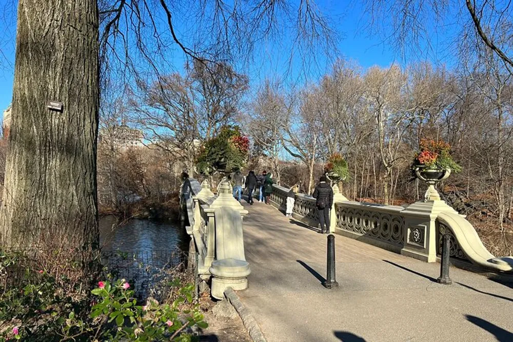 A scenic bridge with ornate railings and flower arrangements crosses over a tranquil body of water in a park where people are strolling and appreciating a sunny day