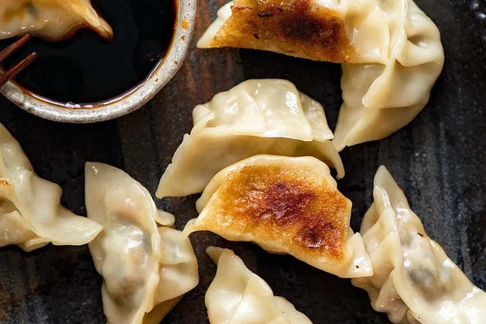 The image shows several golden-brown pan-fried dumplings next to a small bowl of dipping sauce on a dark surface