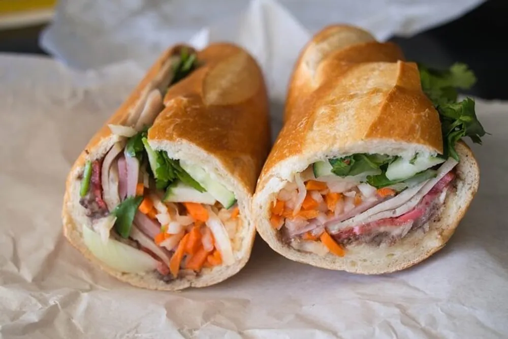 The image shows a freshly cut Vietnamese banh mi sandwich with various fillings including meats pickled vegetables and herbs placed on a piece of parchment paper