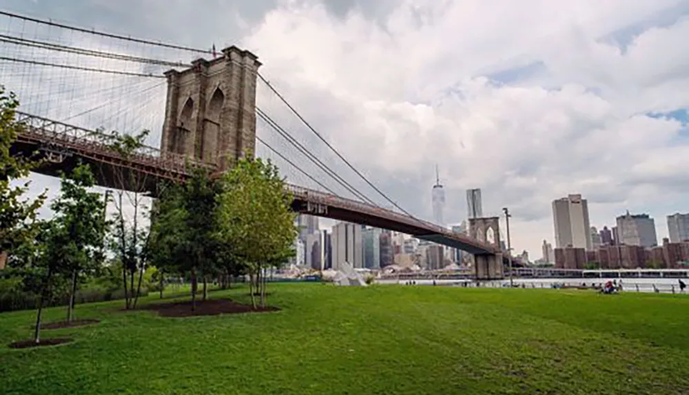 The Brooklyn Bridge spans across the East River with the Manhattan skyline in the background viewed from a grassy park area