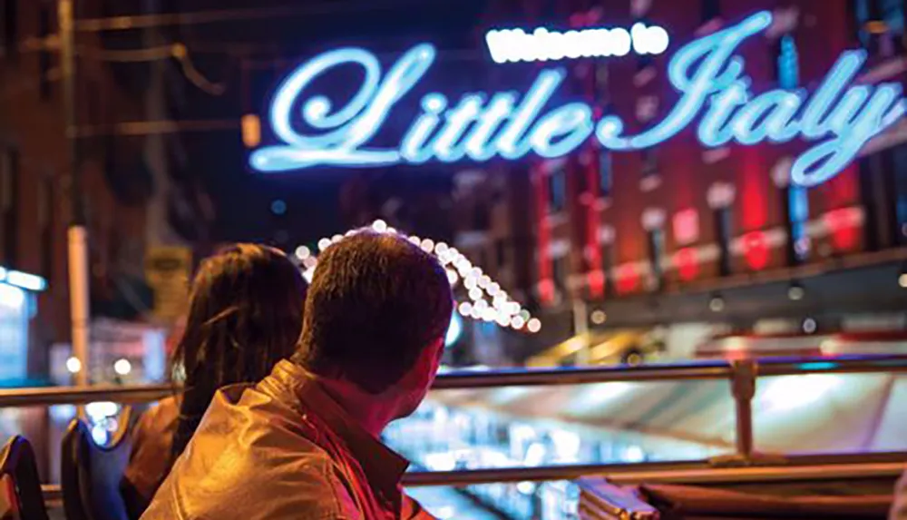 Two people are sitting and looking towards a neon sign that reads Welcome to Little Italy suggesting an intimate urban scene at night