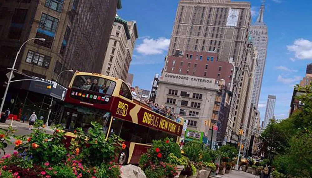 A sightseeing bus tours through a vibrant urban street lined with towering buildings and lush flowers under a blue sky