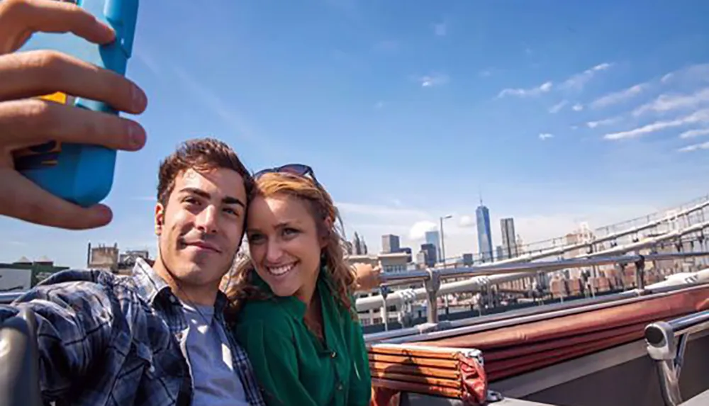 A smiling couple is taking a selfie with a city skyline including a tall skyscraper in the background