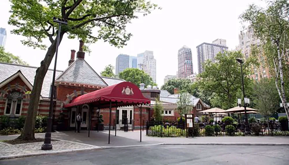 The image shows a quaint red-brick building with a red awning set in a serene urban park surrounded by lush greenery and tall buildings looming in the background