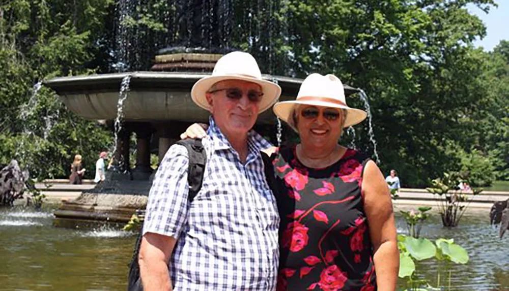 A smiling couple wearing hats stands in front of a water fountain on a sunny day
