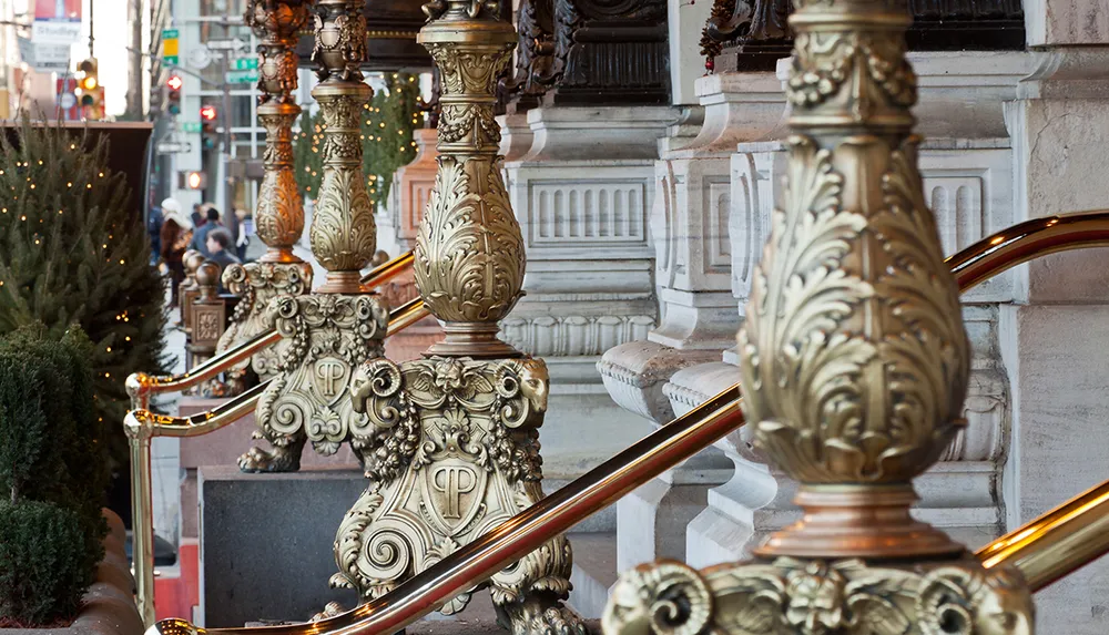 The image shows an ornately decorated staircase with polished brass railings and intricate gilded posts set against an urban street background with pedestrians