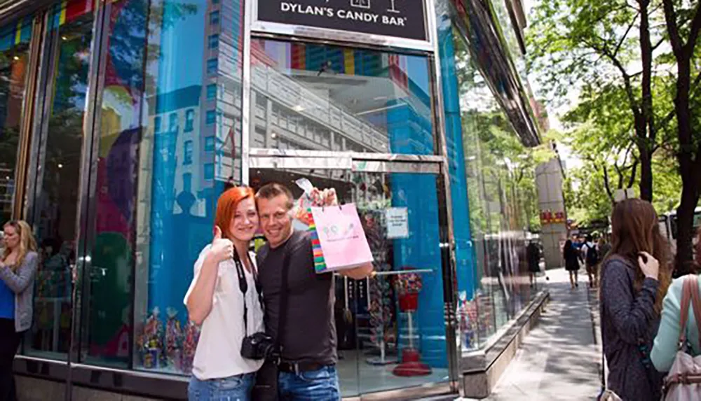 A happy couple poses with a shopping bag in front of Dylans Candy Bar surrounded by vivid colors and city life