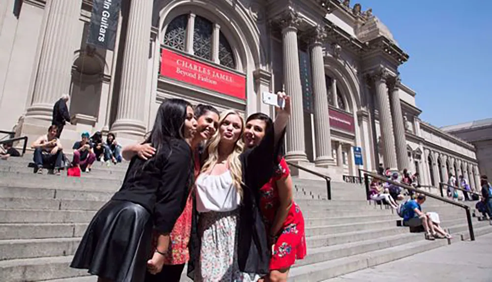 A group of friends is taking a selfie together on the steps of a grand building with classical architecture under a bright blue sky