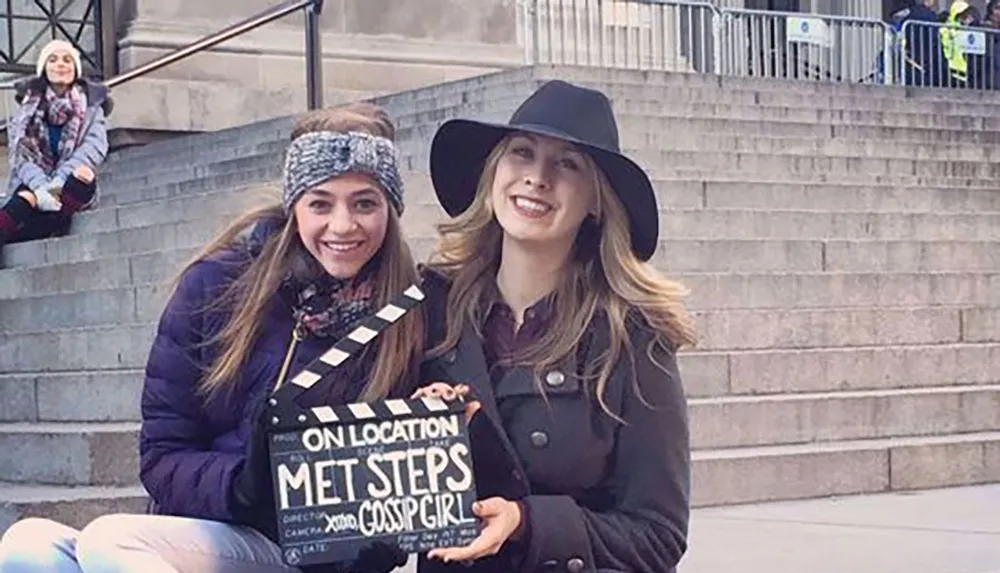 Two smiling women pose for a photo on some steps with one holding a clapperboard labeled MET STEPS GOSSIP GIRL
