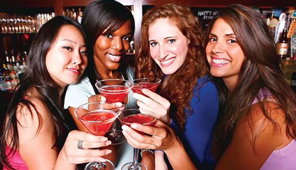 Four smiling women are holding and toasting with red cocktails at a bar