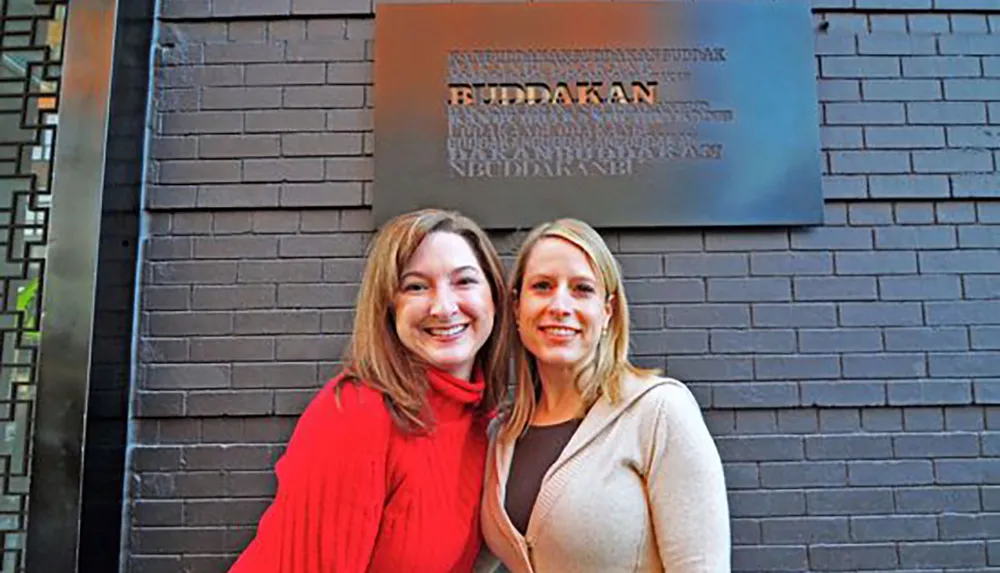 Two smiling women are posing together in front of the entrance of a location named Buddakan