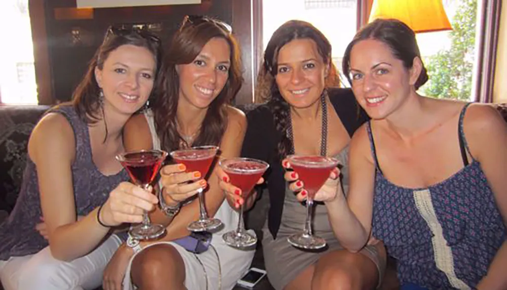 Four smiling individuals are sitting closely together toasting with red cocktails in a cozy indoor setting