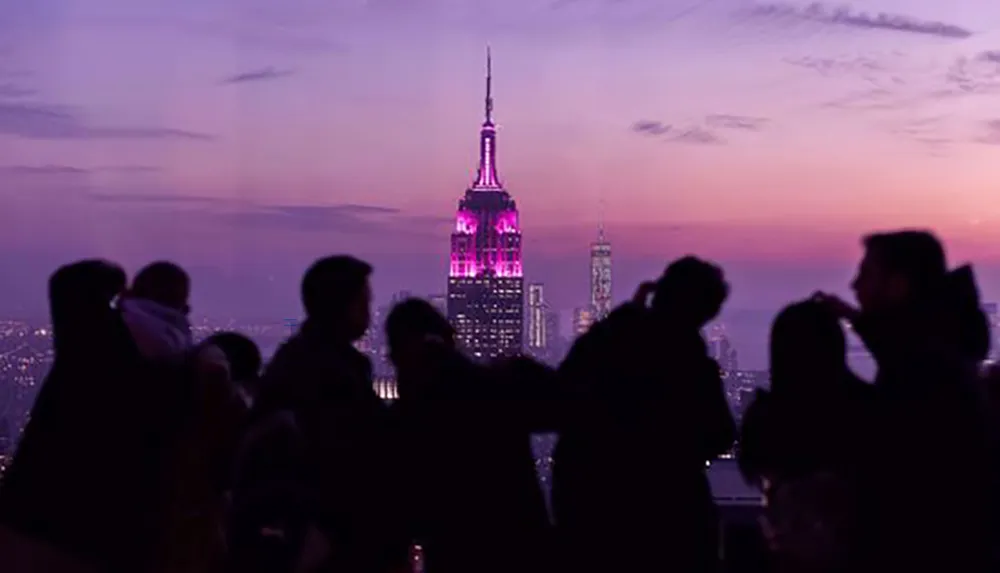 People are gathered at a high vantage point during dusk enjoying the view of a city skyline punctuated by a prominently illuminated building under a gradient twilight sky