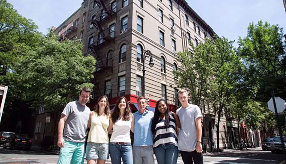 A group of six people is posing and smiling on a sunny street in front of a historic building with a brick facade
