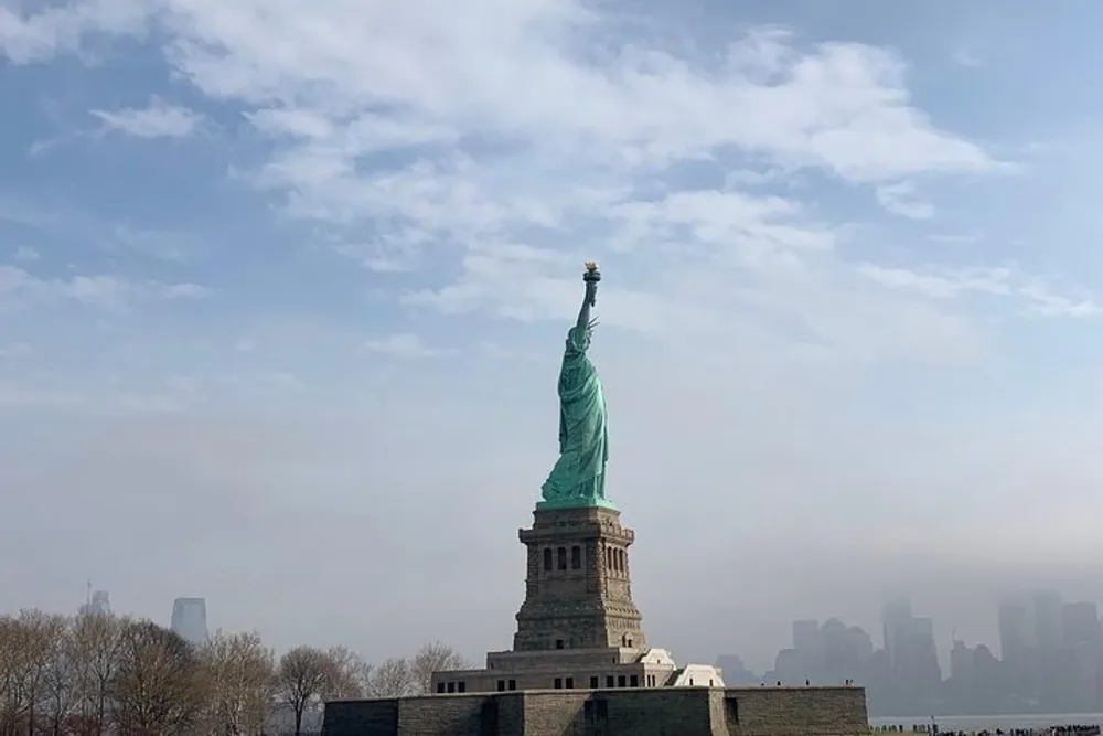 The Statue of Liberty stands tall with a clear sky above and a faint city skyline in the background likely representing its iconic presence as a symbol of freedom and welcome to the United States