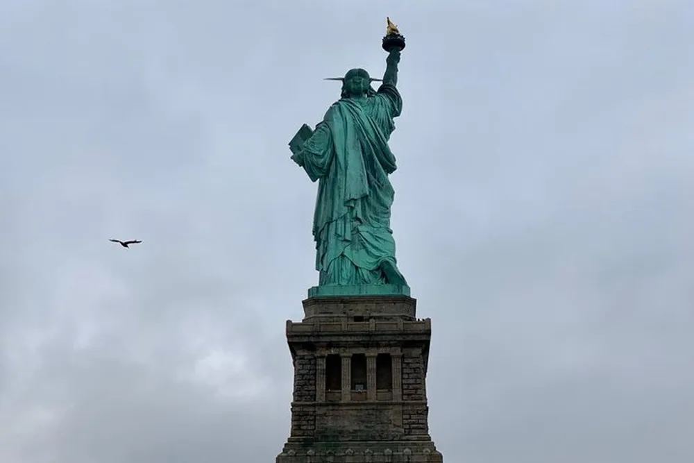 The image shows the Statue of Liberty against a cloudy sky with a bird flying on the left side
