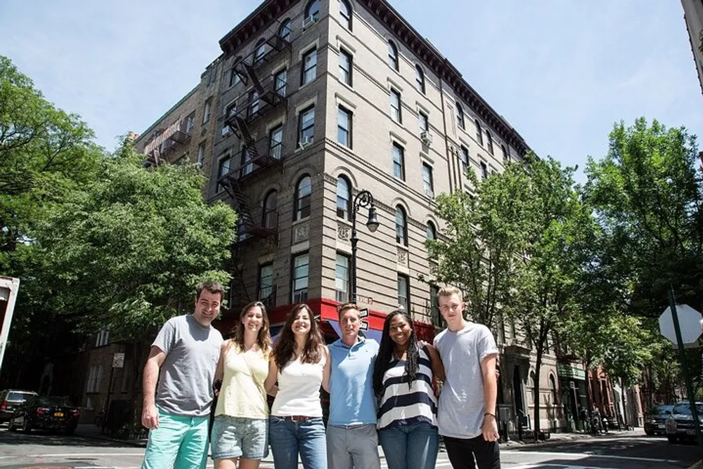 A group of seven individuals is smiling for the camera in front of an old corner building on a sunny day with trees and parked cars along the street