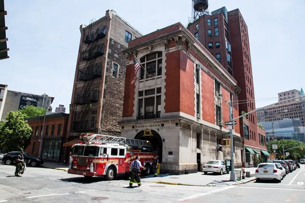 A fire truck is parked outside a red brick fire station with firefighters walking around on a clear day