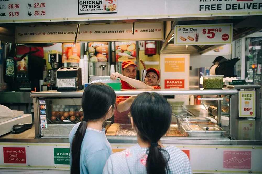 Two individuals are looking at a fast food stall where workers in red and yellow uniforms appear to be engaging with them from behind the counter offering various food items such as hot dogs and drinks