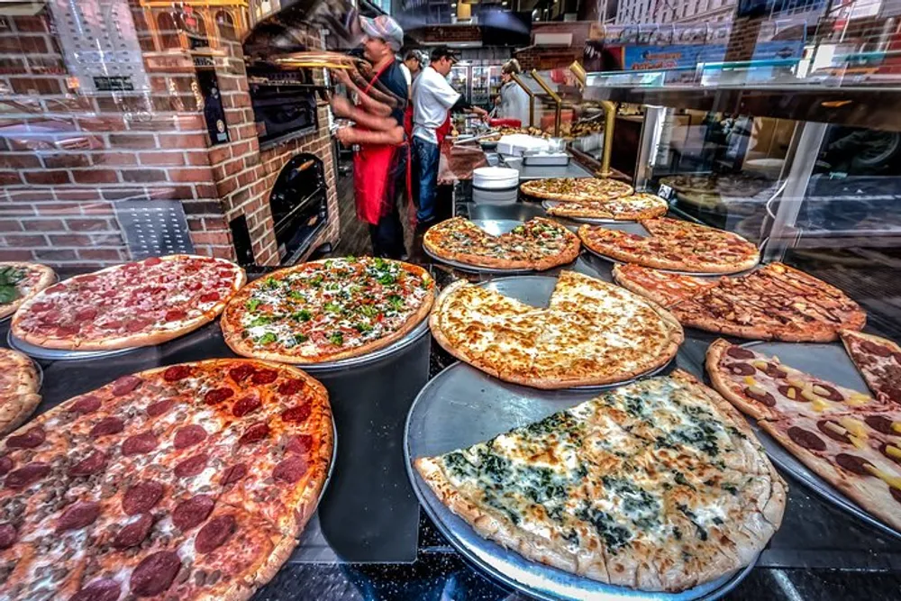 A selection of large freshly prepared pizzas is displayed in the foreground with chefs working in the background at a pizzeria