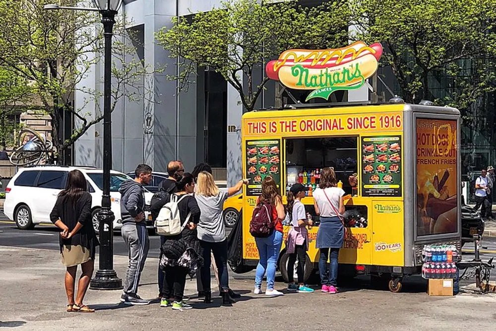 A group of people are standing in line to order food from a vibrant yellow Nathans hot dog cart on a sunny day in an urban setting
