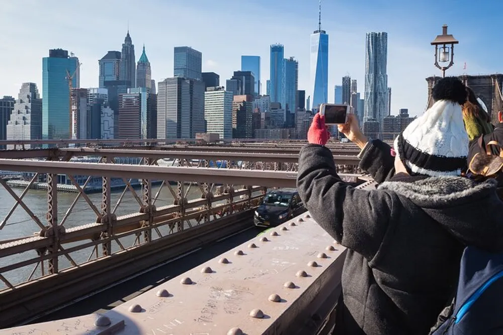 A person wearing a hat is taking a photo of a city skyline with a smartphone from a bridge on a sunny day