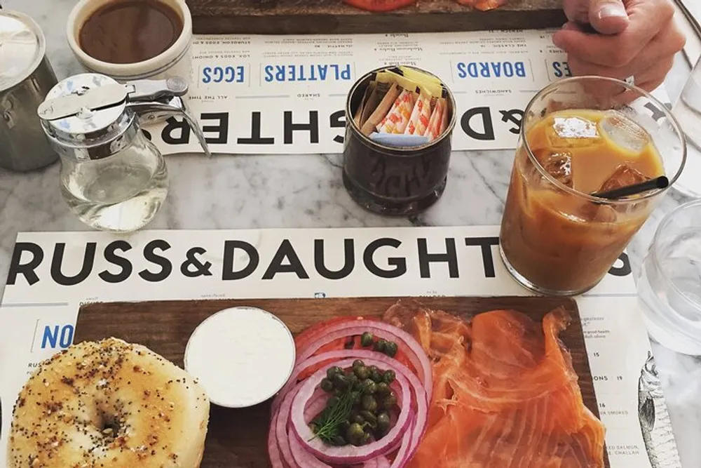 This image features a table setting with bagels smoked salmon cream cheese onions capers an iced coffee and a hot beverage likely at Russ  Daughters caf