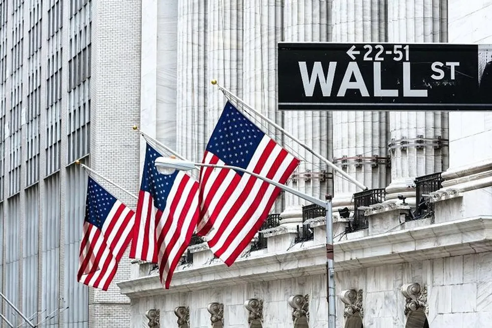 The image shows several American flags fluttering in front of a building beneath a street sign that reads 22-51 Wall St indicating the location is Wall Street